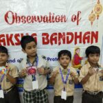 Four schoolboys in beige uniforms display handmade rakhi crafts in front of a banner that reads observation of raksha bandhan